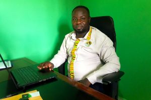 kiwanuka-musa-lubega Man sitting at a desk with a laptop in front of a bright green screen background, wearing a light-colored shirt with colorful trim.